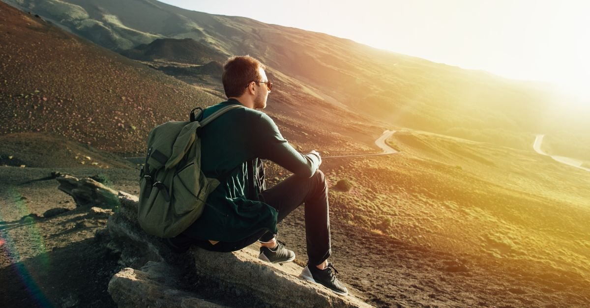 A traveler with a backpack sitting on a rock, gazing at the mountain landscape during sunset.
