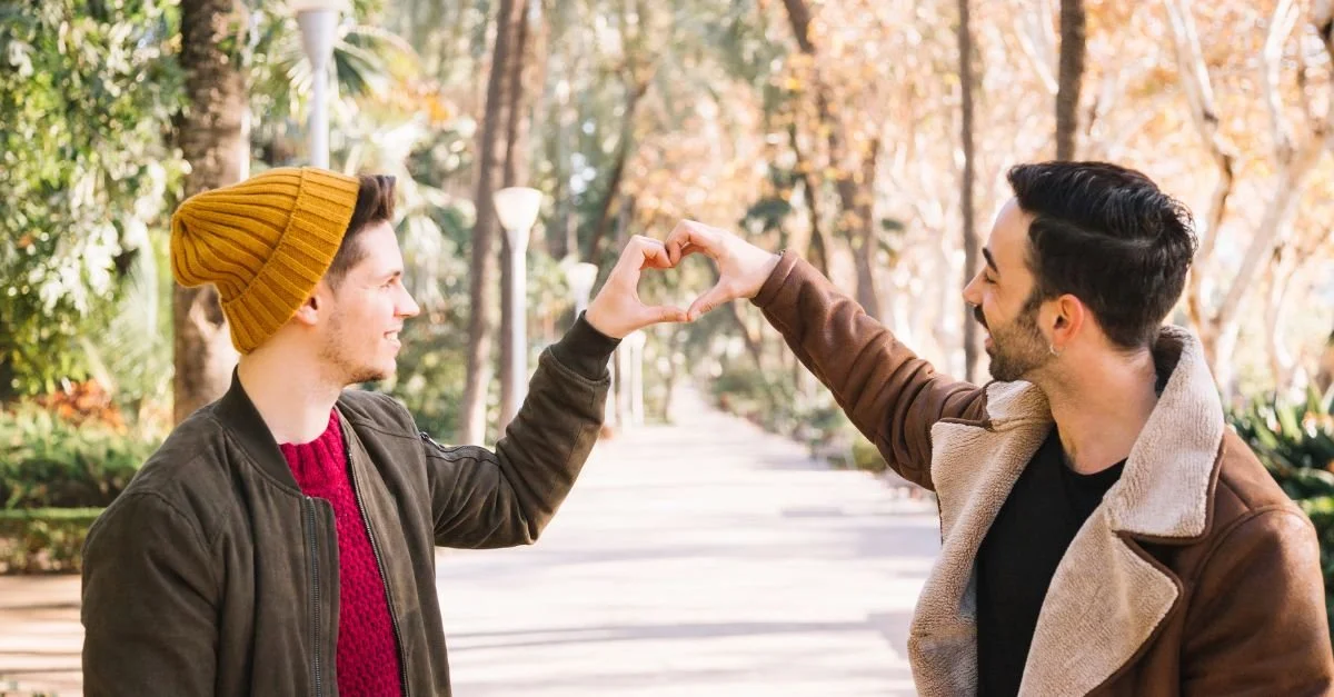 Two friends smiling and creating a heart shape with their hands while standing on a path surrounded by trees.